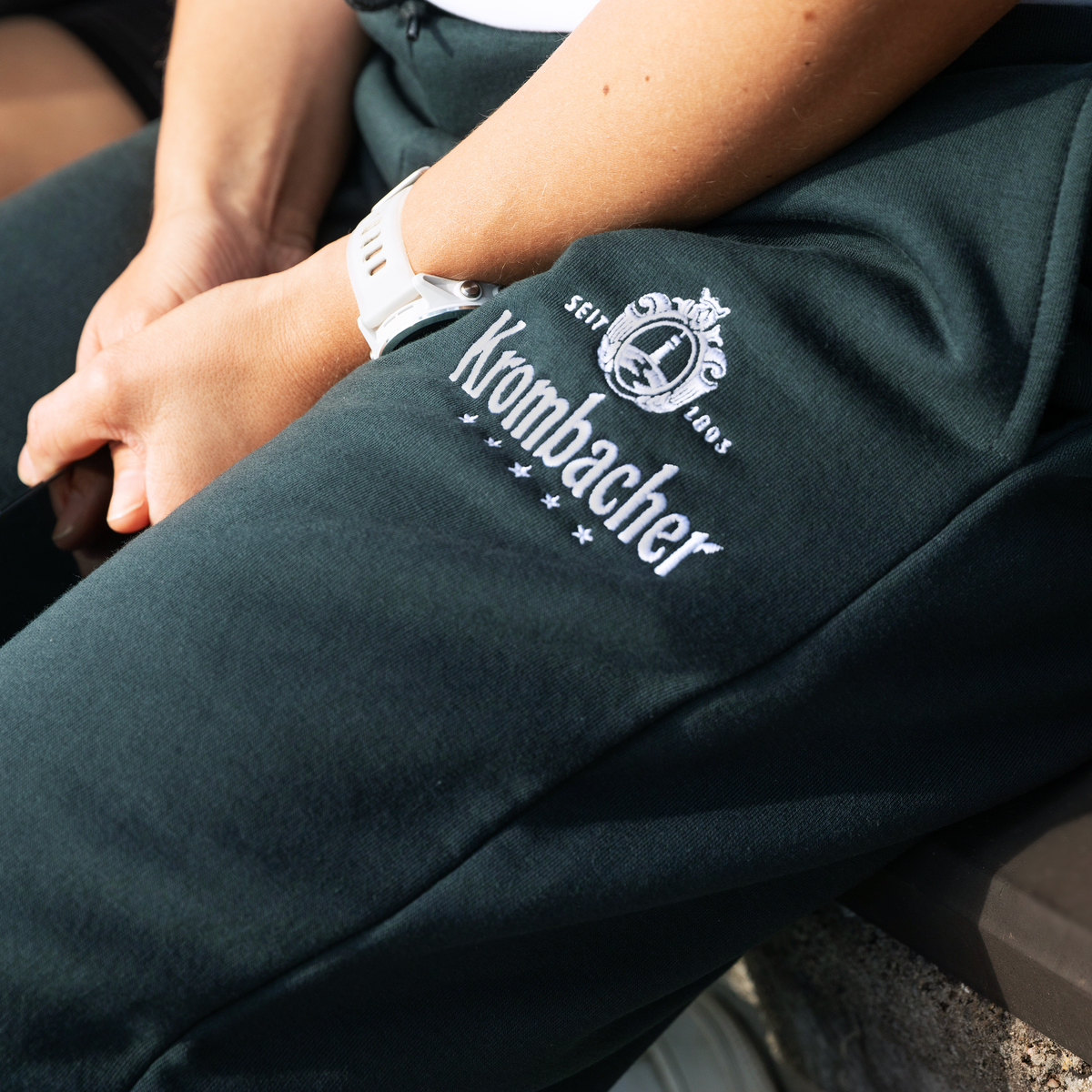 Nahaufnahme eines dunkelgrünen Jogginghosenbeins mit weißem Krombacher-Logo und Schriftzug sowie einer weißen Armbanduhr am Handgelenk einer verschränkten Hand