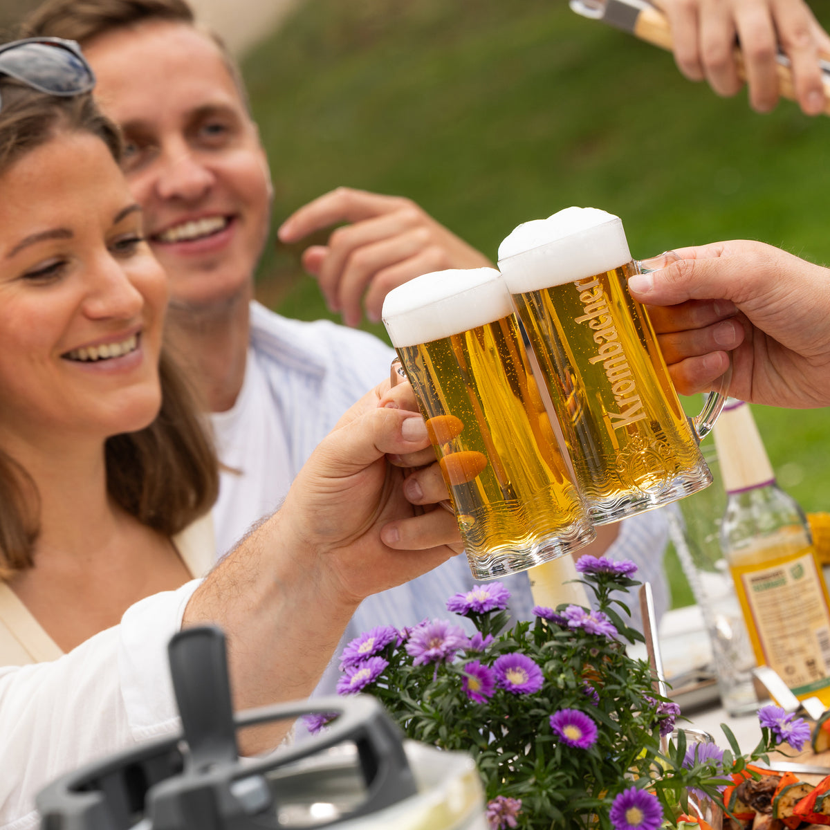 Drei Personen stoßen mit Biergläsern mit Schaumkrone an, im Vordergrund violette Blüten auf einem Tisch