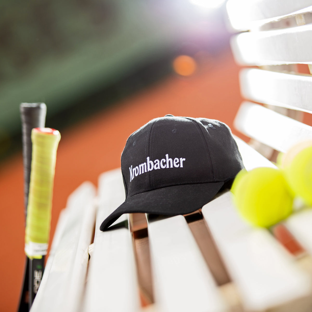 Sportliche schwarze Baseballkappe mit Krombacher Schriftzug auf einer Bank am Tennisplatz.