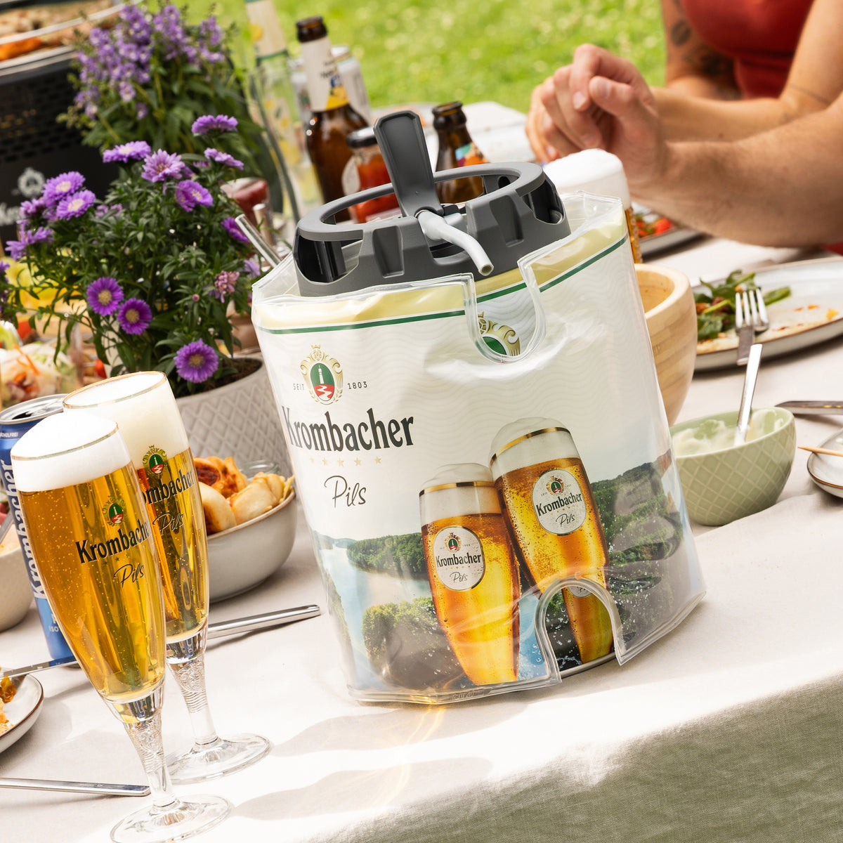 Krombacher-Pils-Eispack mit Flaschen und Gläsern auf gedecktem Gartentisch neben lila Blumen und Personenhand