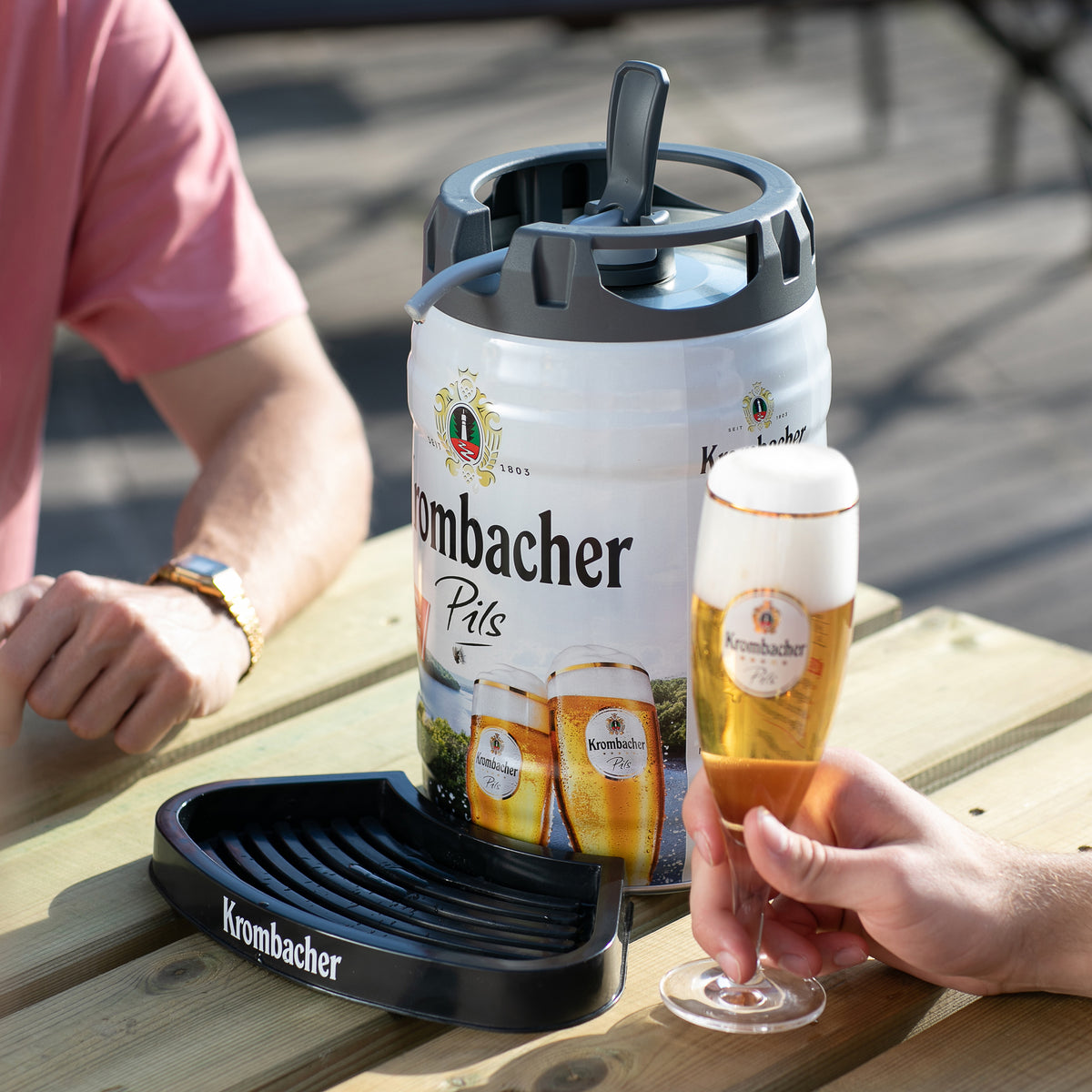 Krombacher-Pils-Fass mit Tropfschale auf Holztisch, eine Hand hält ein gefülltes Krombacher-Glas, eine Person mit rosa Hemd sitzt daneben