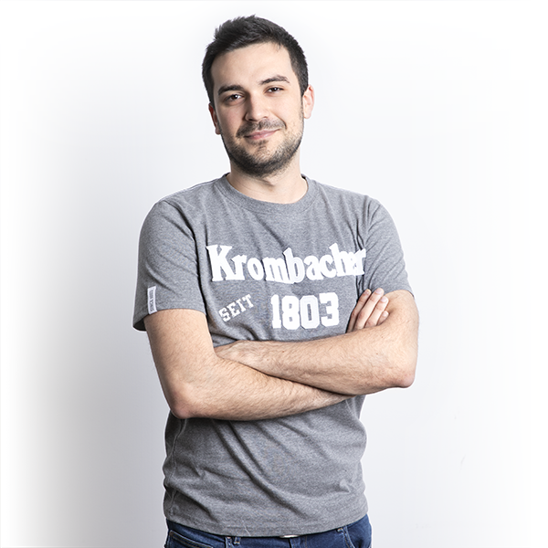 Mann mit verschränkten Armen trägt graues T-Shirt mit weißem Schriftzug 'Krombacher seit 1803'.