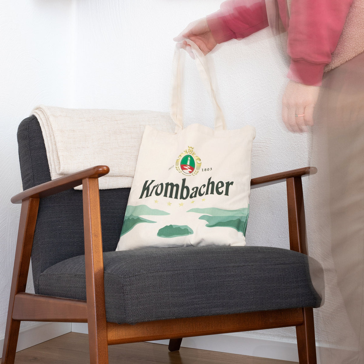 Beige Jutebeutel mit Krombacher-Logo, See und Schriftzug auf dunkelgrauem Sessel mit Holzarmlehnen, Person hält Henkel.