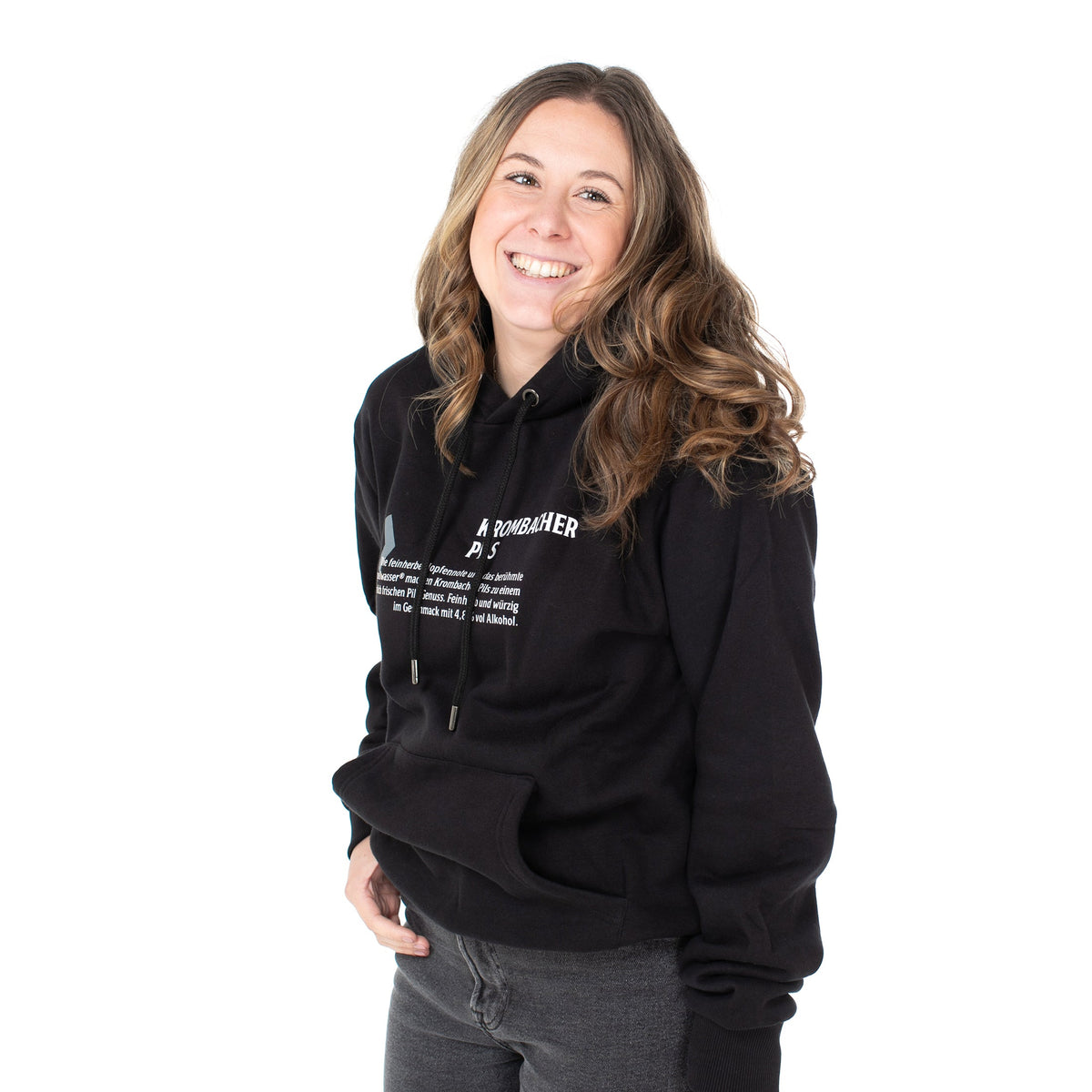 Person trägt schwarzen Hoodie mit weißem Text 'Krombacher Pils' und Kapuze, kombiniert mit grauer Jeans.