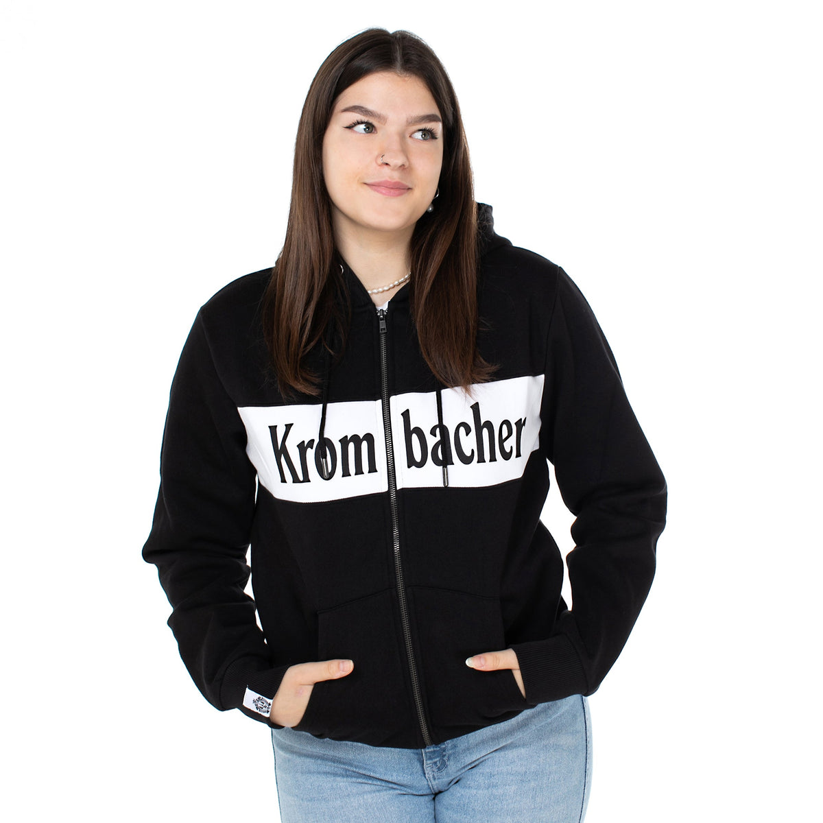 Person trägt schwarze Sweatshirtjacke mit weißem Schriftzug 'Krombacher' auf der Brust, Hände in den Taschen.