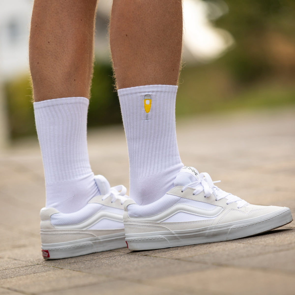 Weiße Sportsocken mit kleinem Bierglas-Logo, getragen mit weißen Sneakern auf gepflastertem Boden.
