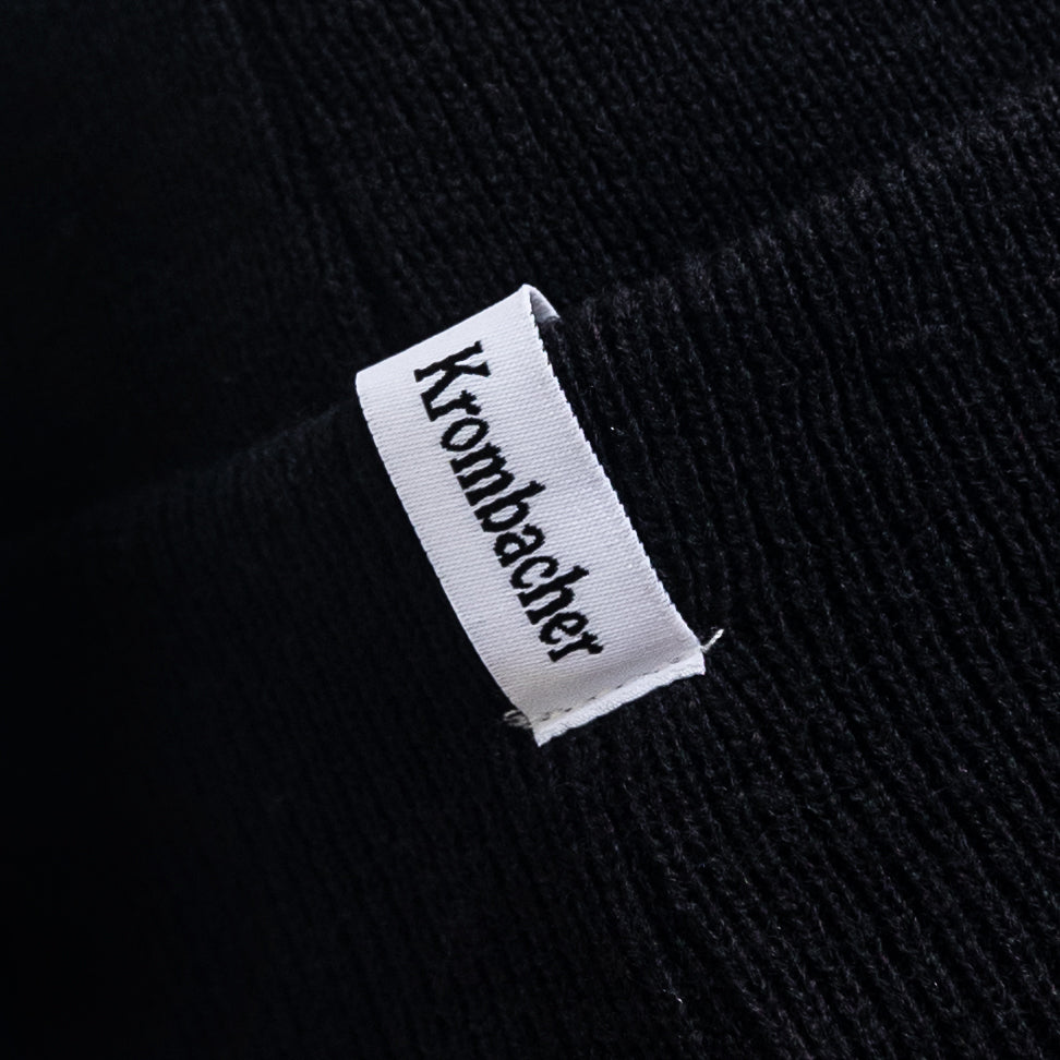 Schwarze Beanie mit weißem Etikett und schwarzem Schriftzug 'Krombacher'.