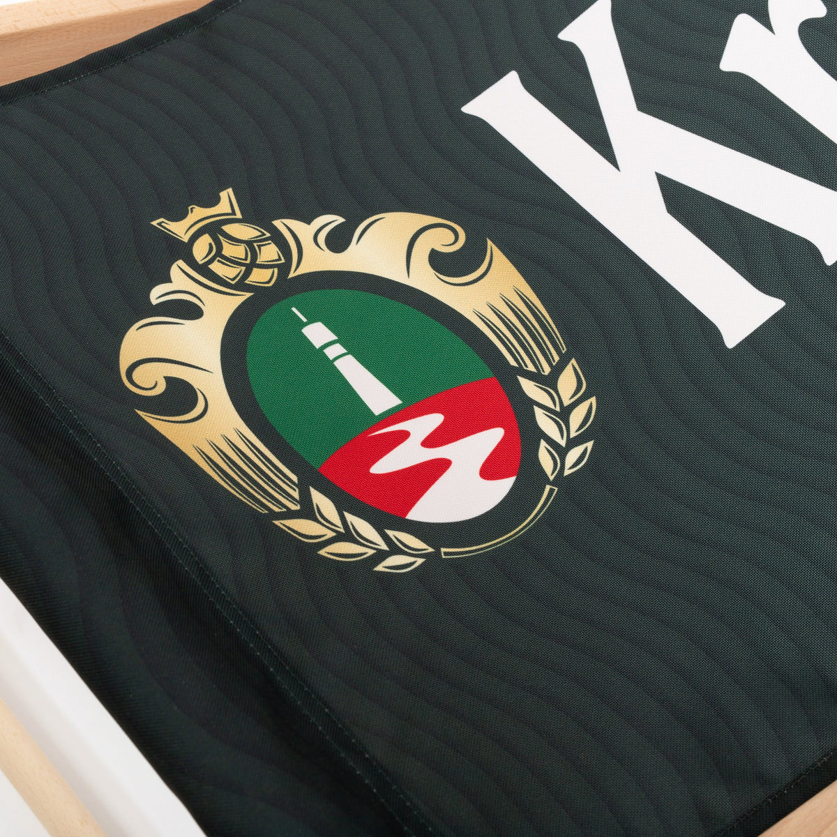 Detail des Krombacher-Logos mit grünem und rotem Wappen auf dunkelgrünem Stoff eines Liegestuhls.