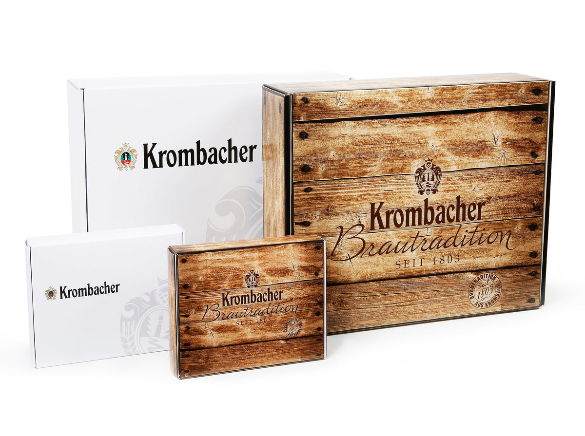 Vier Krombacher Geschenkboxen in verschiedenen Größen, zwei mit Holzoptik und Aufschrift 'Krombacher Brautradition seit 1803', zwei weiße mit Krombacher-Logo.