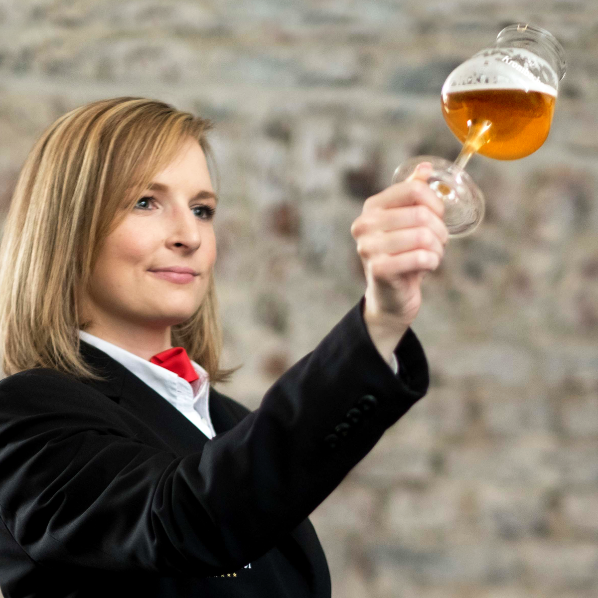 Frau in schwarzem Sakko und rotem Halstuch hält ein Bier gefülltes Sommelierglas vor einem Steinmauerhintergrund.
