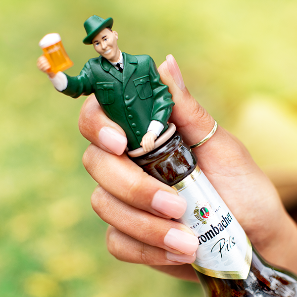 Hand hält Flasche Krombacher Pils mit grünem Flaschenaufsatz in Form eines Försters, der ein Bierglas hebt.