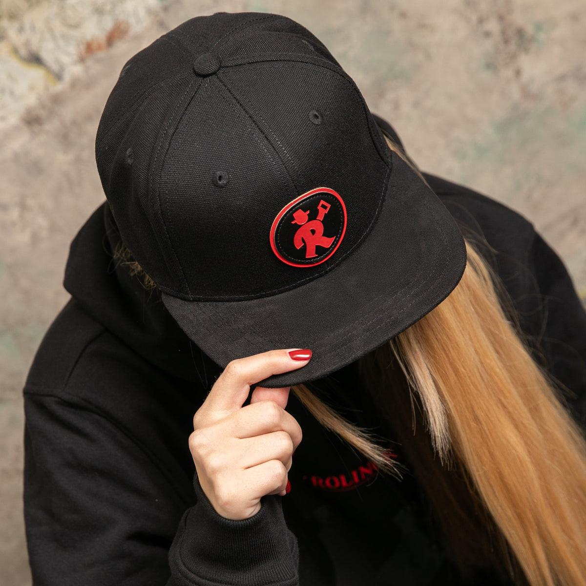Schwarze Cap mit rundem roten Rolinck Logo, getragen von Person mit langen blonden Haaren und rotem Nagellack.
