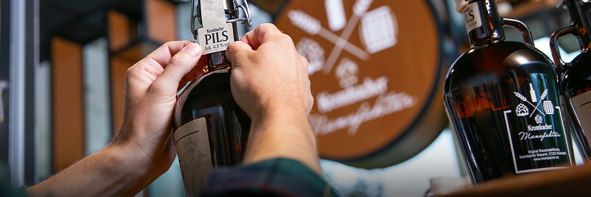 Person befestigt Etikett mit Aufschrift 'Krombacher Pils' an brauner Glasflasche in Krombacher Manufaktur