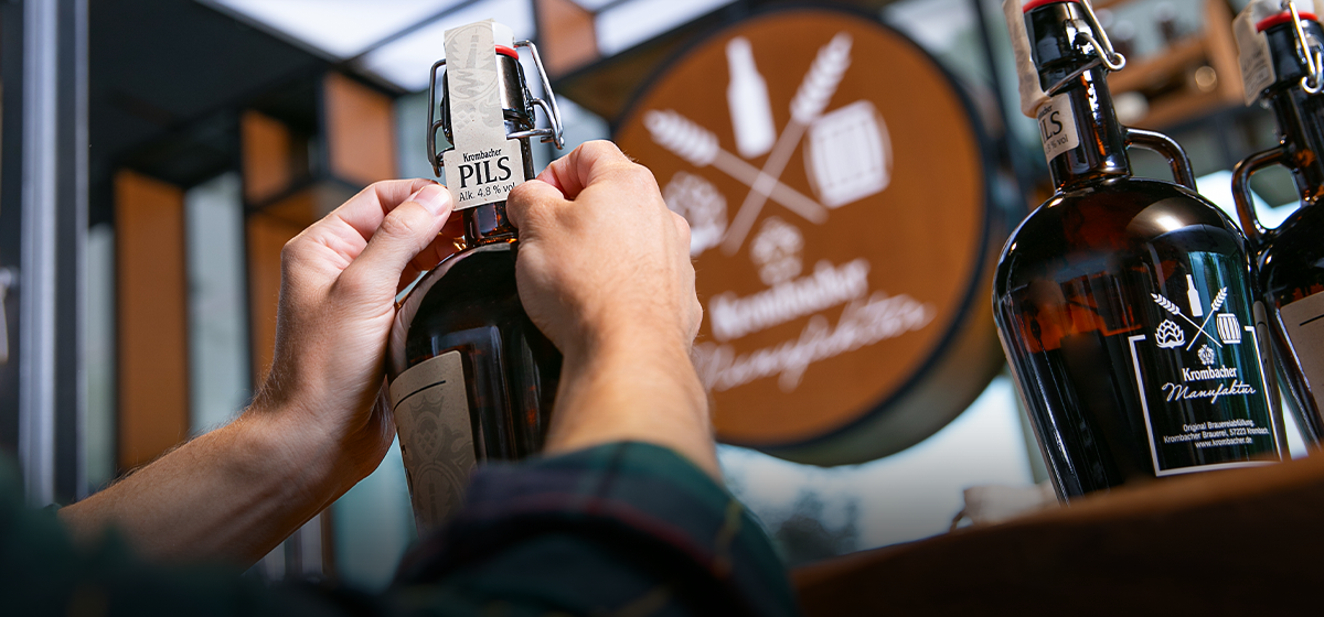Person befestigt Etikett mit Aufschrift 'Krombacher Pils' an brauner Glasflasche in Krombacher Manufaktur