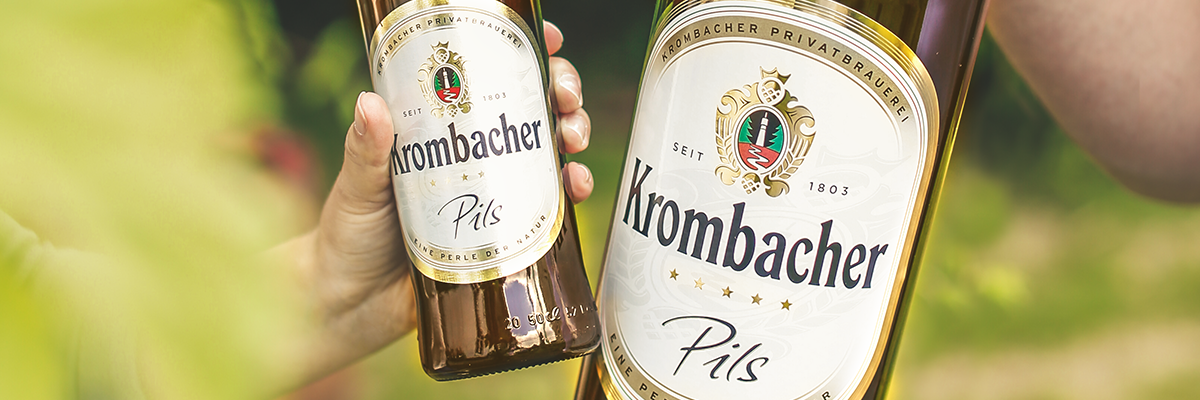 Zwei Hände halten Krombacher Pils Bierflaschen, eine 3-Liter-Flasche und eine Standardflasche, im Freien