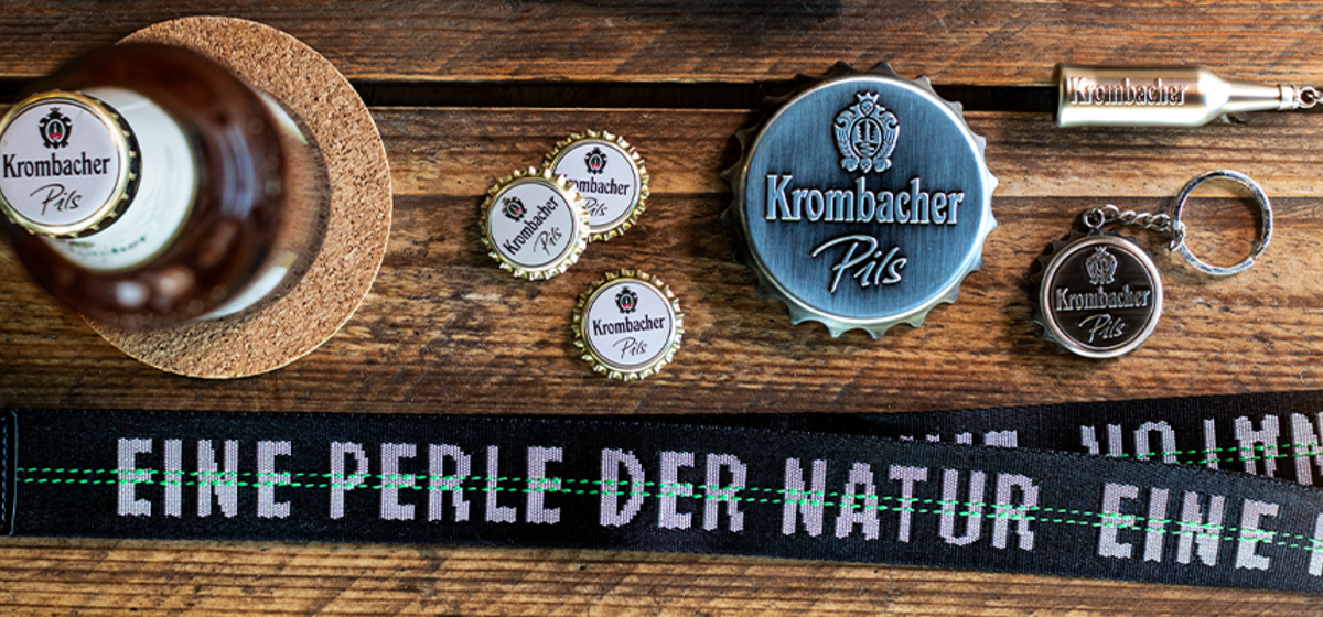 Krombacher Pils Bierflasche, Kronkorken, Flaschenöffner und Schlüsselanhänger mit Krombacher-Logo, Text auf Band: 'EINE PERLE DER NATUR'