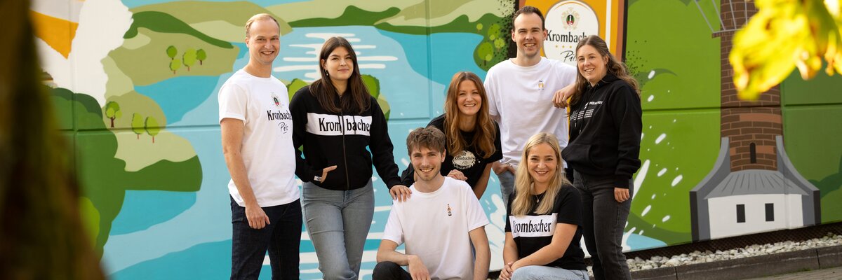 Sieben junge Erwachsene tragen Krombacher T-Shirts und Hoodies vor einer bunten Wand mit Landschaftsmotiv