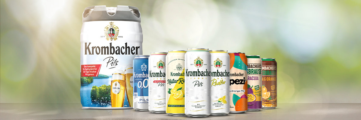 Verschiedene Krombacher Bier- und Erfrischungsgetränkedosen und ein 5-Liter-Fass Krombacher Pils nebeneinander auf Holzoberfläche