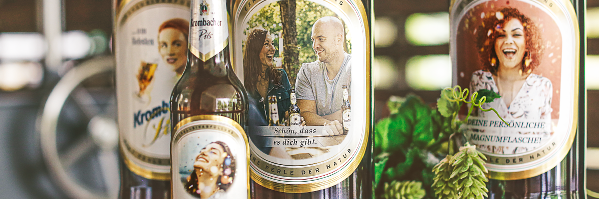 Drei Krombacher Bierflaschen in verschiedenen Größen mit Etiketten, die Menschen und Naturmotive zeigen