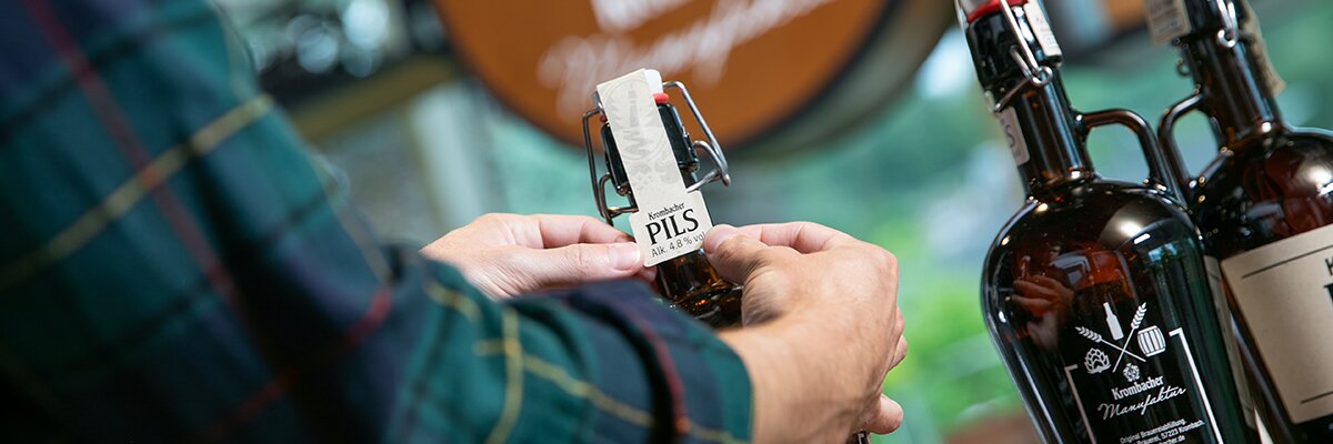 Person beklebt eine Flasche mit Halsetikett 'Krombacher Pils'