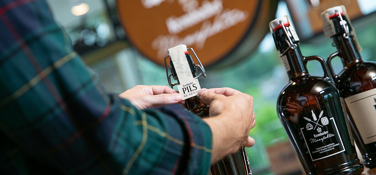 Person beklebt eine Flasche mit Halsetikett 'Krombacher Pils'