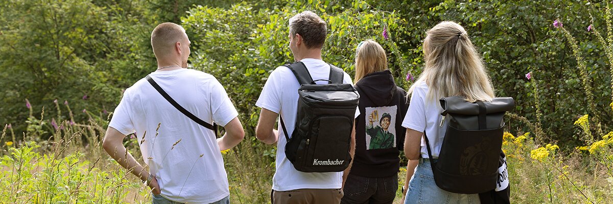 Vier Personen mit Rucksäcken und weißen T-Shirts gehen auf einem Weg durch eine grüne Wiese mit Bäumen