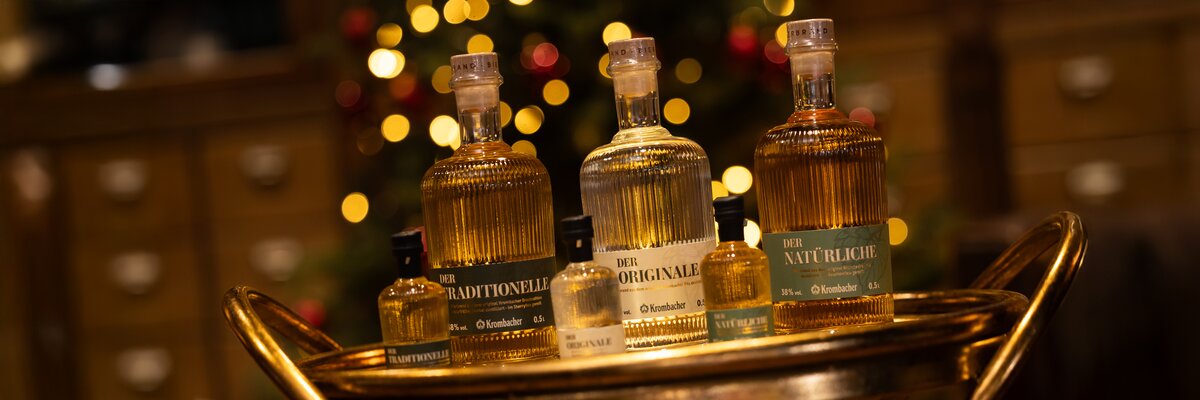 Mehrere unterschiedlich große Glasflaschen mit Krombacher Bierbrand Inhalt stehen auf einem goldfarbenen Tablett vor unscharfem Hintergrund mit Lichterketten.