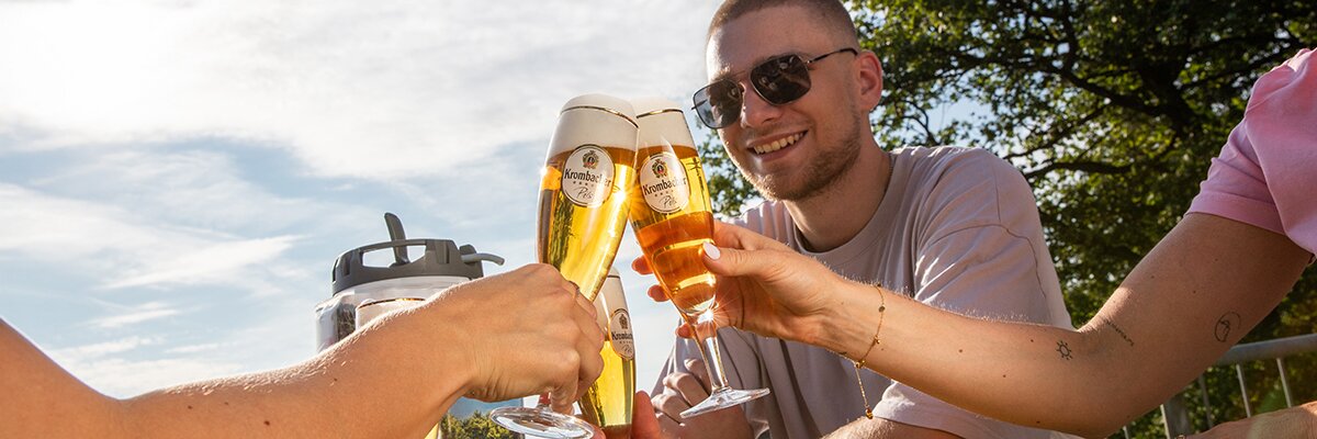 Drei Personen stoßen mit gefüllten Krombacher Biergläsern im Freien an, sonniger Himmel und Bäume im Hintergrund