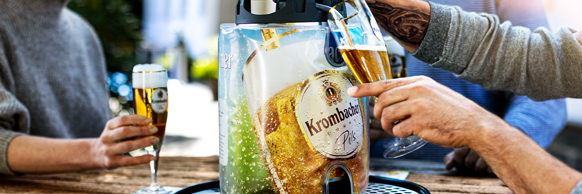 Personen zapfen Krombacher Pils aus einem Frischefässchen in Gläser auf einem Holztisch im Freien