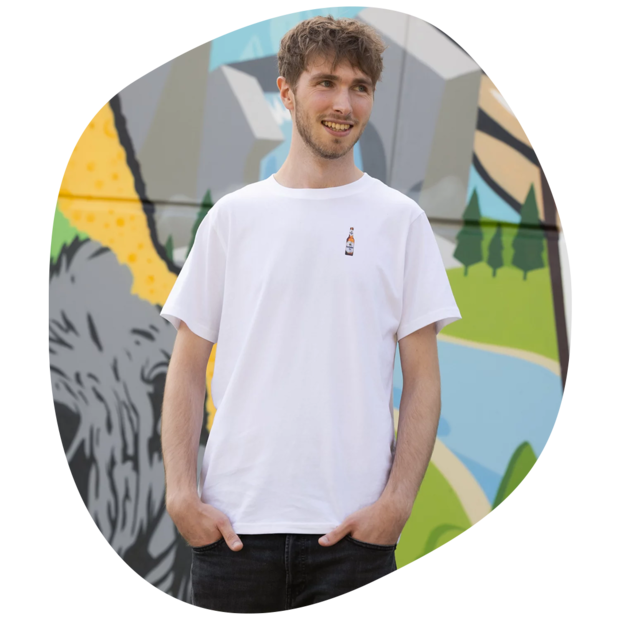 Mann trägt weißes T-Shirt mit kleinem Flaschenmotiv auf der Brust, steht vor bunter Wand mit Graffiti