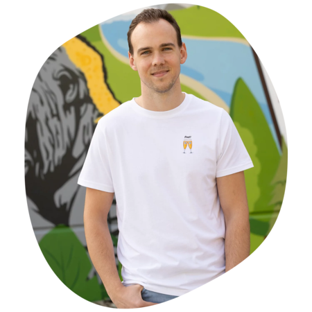 Mann trägt weißes T-Shirt mit kleinem Aufdruck von zwei anstoßenden Biergläsern und dem Wort 'Prost' vor bunter Wand mit Graffiti