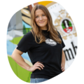 Frau mit langen Haaren, trägt schwarzes T-Shirt mit rundem weißen Kronkorken-Logo und blaue Jeans, steht mit Hand in der Hüfte vor bunter Wand