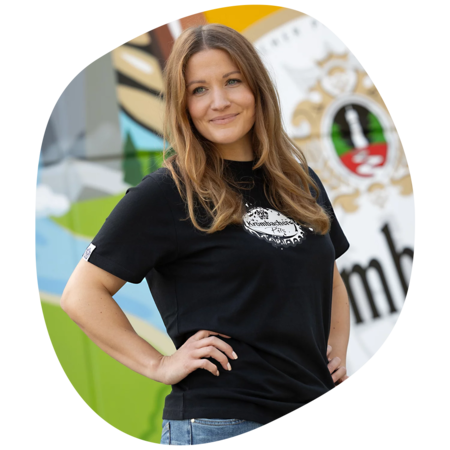 Frau mit langen Haaren, trägt schwarzes T-Shirt mit rundem weißen Kronkorken-Logo und blaue Jeans, steht mit Hand in der Hüfte vor bunter Wand