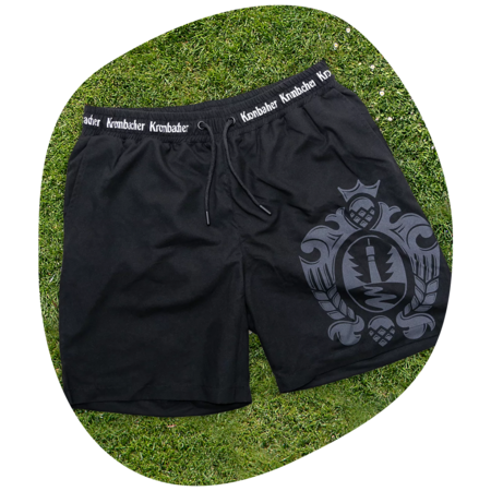 Schwarze Badeshorts mit Kordelzug und weißem Schriftzug 'Krombacher' am Bund, großes Wappenlogo auf dem linken Bein, liegend auf Gras