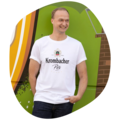 Mann trägt weißes T-Shirt mit schwarzem Schriftzug 'Krombacher Pils' und Wappen, steht vor grüner Wand mit Graffiti