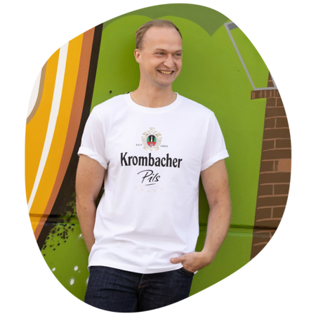 Mann trägt weißes T-Shirt mit schwarzem Schriftzug 'Krombacher Pils' und Wappen, steht vor grüner Wand mit Graffiti