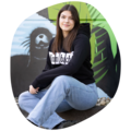 Frau sitzt im Schneidersitz vor einer Wand mit einem Graffiti, trägt schwarze Sweatshirtjacke mit weißem Schriftzug 'Krombacher' und blaue Jeans