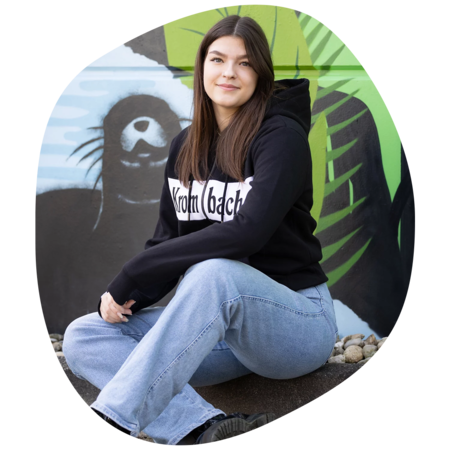 Frau sitzt im Schneidersitz vor einer Wand mit einem Graffiti, trägt schwarze Sweatshirtjacke mit weißem Schriftzug 'Krombacher' und blaue Jeans