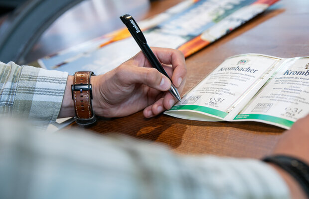 Hand mit Stift beschriftet Krombacher Rückenetikett mit dem Mindesthaltbarkeitsdatum auf Holztisch, Person trägt kariertes Hemd und Lederarmband