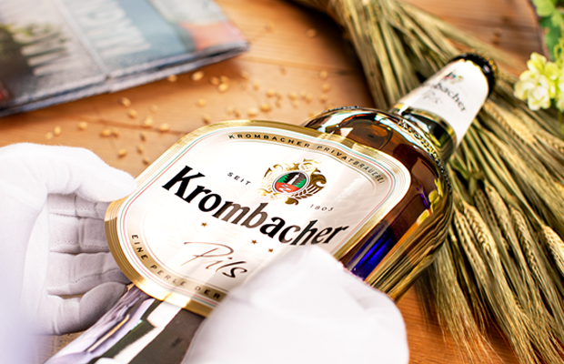 Hände mit weißen Handschuhen beklebt eine Krombacher Pils 3,0l Flasche mit einem Bauchetikett, auf dem das Krombacher Logo abgebildet ist