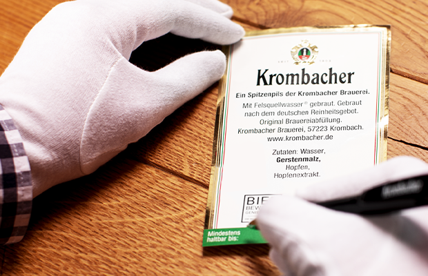 Hände mit weißen Handschuhen beschreibt ein Krombacher Rückenetikett mit dem Mindesthaltbarkeitsdatum