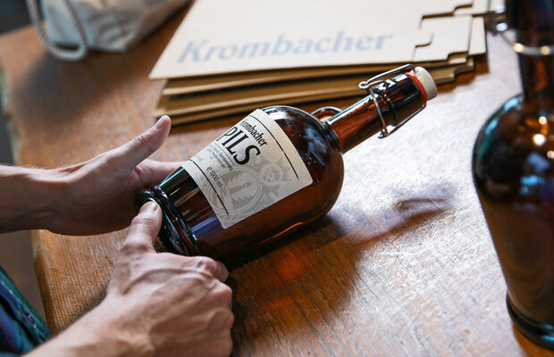 Person bringt Etikett mit Schrift 'Krombacher Pils' auf braune Bierflasche mit Bügelverschluss an