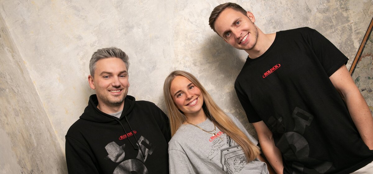 Drei Personen stehen vor einer hellen Wand, zwei tragen schwarze T-Shirts mit roten Schriftzügen, eine trägt ein graues T-Shirt mit schwarzer Rolinck Brauerei.