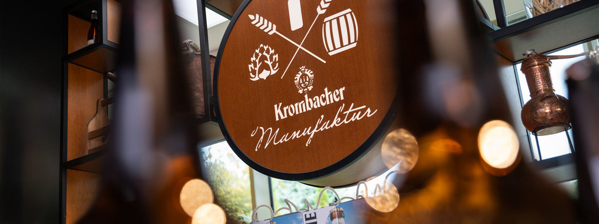 Krombacher Manufaktur im Ausstellungsraum mit Holzschild, Brausymbolen und Flaschen im Vordergrund.