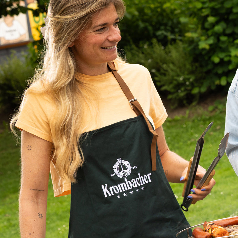 Krombacher Grill-Bundle mit Grillschürze und Grillzange bei einer Gartengrill-Szene.