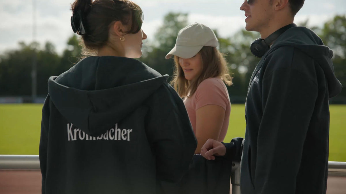 Drei Personen in Krombacher-Hoodies stehen an einem Sportplatz.
