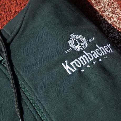 Nahaufnahme eines dunkelgrünen Kapuzenpullovers mit Reißverschluss und weißem Krombacher-Logo