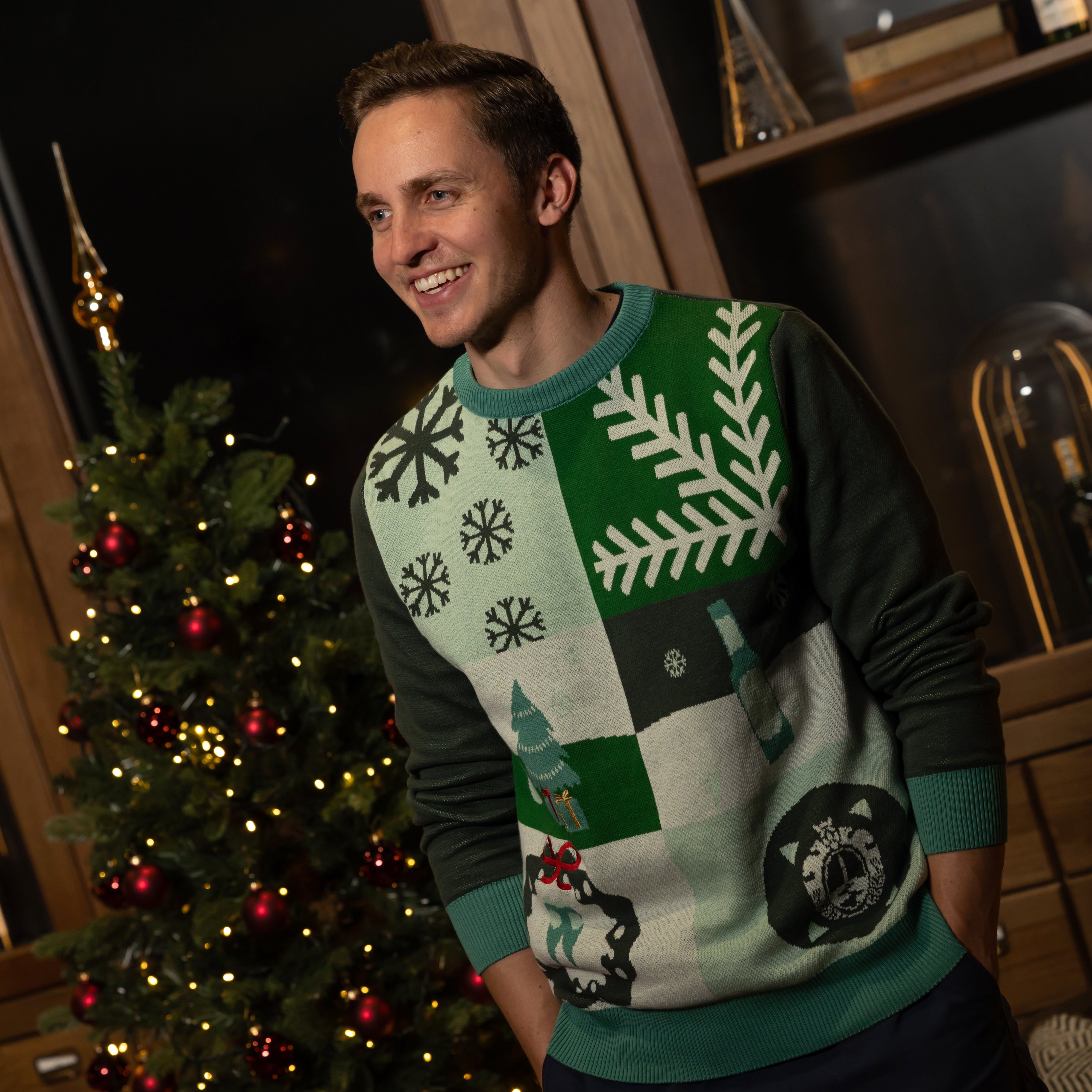 Mann trägt grünen Weihnachtspullover mit Schneeflocken, Tannenbaum und Rentier, steht vor geschmücktem Weihnachtsbaum.