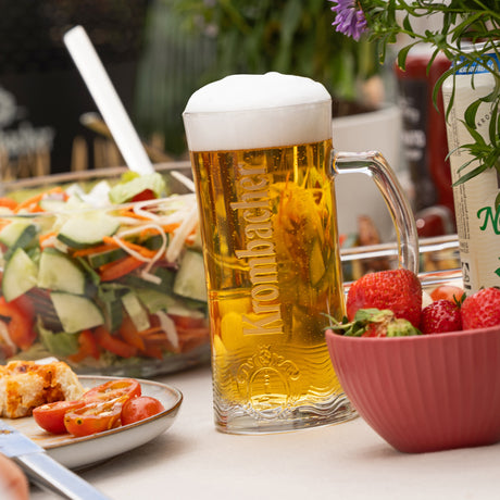 Glas mit Krombacher-Bier mit Schaum neben Schüssel mit Erdbeeren und Salat in Glasschüssel auf gedecktem Tisch