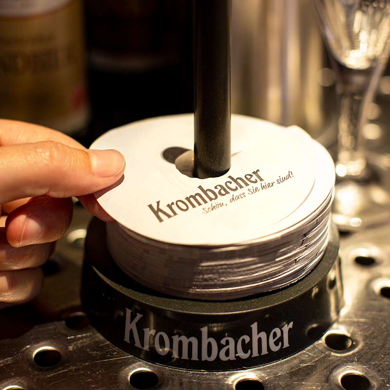 Theken-Szenerie mit einer Hand, die ein Krombacher Tropfendeckchen anhebt.